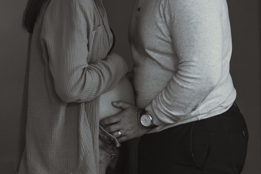 Side profile of a pregnant woman and her husband facing each other with his hands holding her baby bump in a minimalist indoor photoshoot.