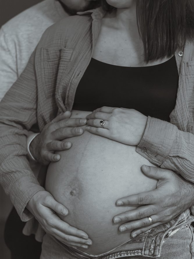 A husband tenderly embracing his pregnant wife from behind, cradling her baby bump in a cozy, black-and-white faceless maternity portrait.